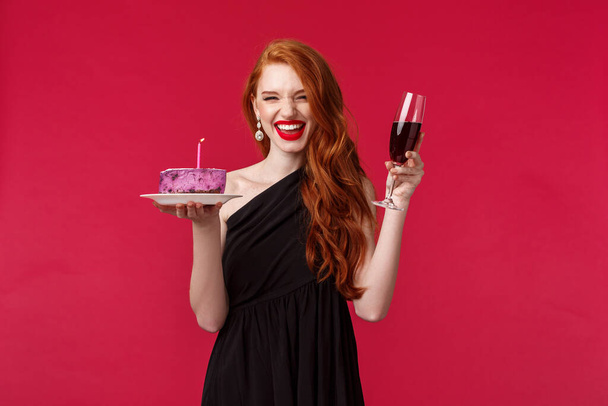 Ritratto di eccitata ridendo splendida rossa che si diverte alla festa di b-day, tenendo un bicchiere di vino e torta di compleanno con candela accesa, esprimendo un desiderio, celebrando su sfondo rosso
 - Foto, immagini