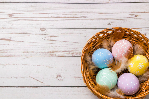 top view of easter eggs hand painted in pastel colors and tender feathers in a wicker basket on wooden table. - Fotó, kép