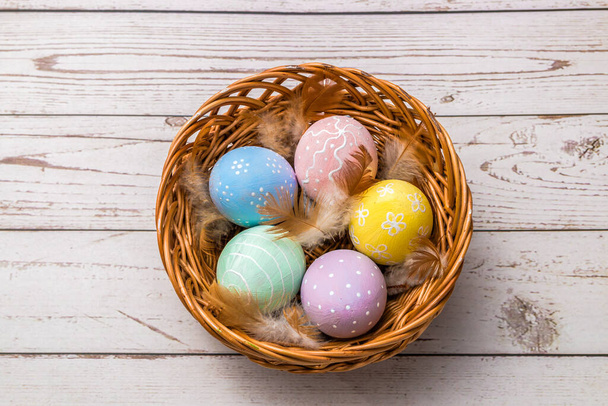 top view of easter eggs hand painted in pastel colors and tender feathers in a wicker basket on wooden table. - Foto, afbeelding