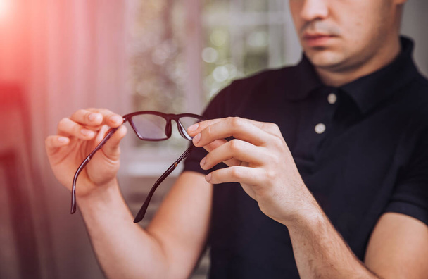 Young man holds stylish black-rimmed eyeglasses. Young male examines glasses in the room. Close-up - Photo, image