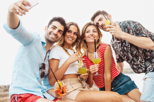 Grupo de jóvenes tomando un selfie con cócteles de aperitivo en la mano contra un cielo blanco. Estilo de vida juvenil concepto de ocio
. - Foto, imagen