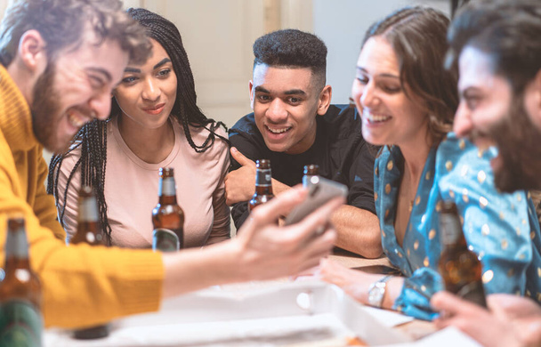 Group of smiling young people watching video on smartphone and drinking beers in a house. Multiracial friends having fun using smartphone, social network apps and new technologies concept. - Photo, Image