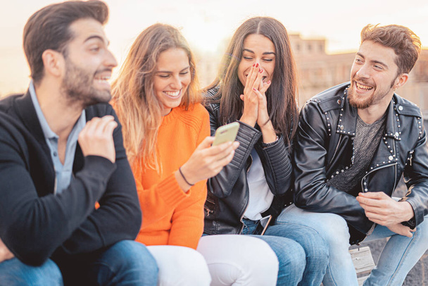 Gruppe von vier Freunden, die zusammen Spaß haben. Zwei Frauen und zwei Männer sitzen lachend auf einer Bank und genießen ihre Zeit mit dem Smartphone. - Foto, Bild