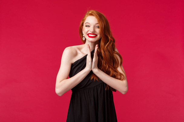 Portrait of happy and pleased cheerful young redhead woman, wearing black formal dress makeup, red lipstick, grinning joyfully, applause as watching fabulous performance, stand red background - Photo, Image
