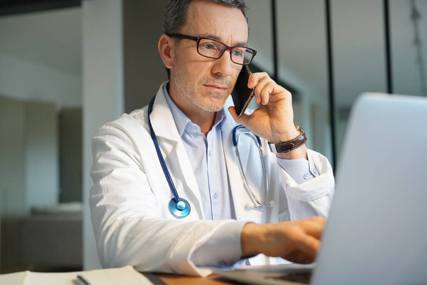 Doctor in office working on laptop talking on phone - Photo, Image