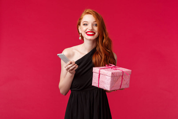 Portrait of happy, excited attractive woman with ginger hair, celebrating birthday, holiday with presents, receive gift, hold box and smartphone laughing smiling camera, red background - Photo, Image