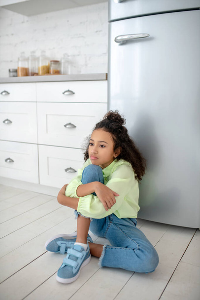 Dark-haired girl in jeans sitting near the fridge feeling hungry - Foto, Imagen