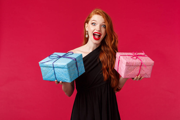 Koncept oslavy, svátků a žen. Portrét vzrušené a šťastné ženské zrzky ženy v černých stylových šatech, držící dva narozeninové dárky, modré a růžové krabice, usmívající se užaslý a veselý - Fotografie, Obrázek
