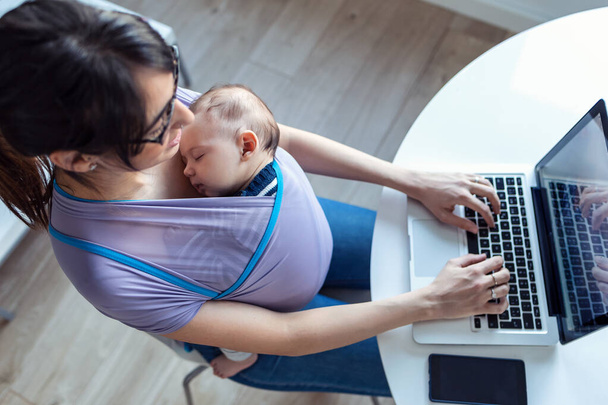 Shot of pretty young mother with her baby in sling working with laptop at home. - Фото, изображение