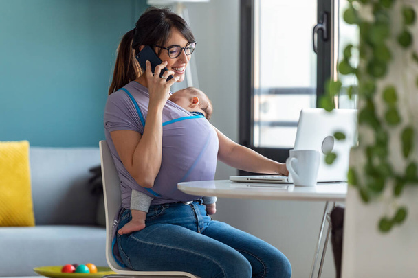 Shot of pretty young mother with her baby in sling talking on mobile phone while working with laptop at home. - Foto, immagini