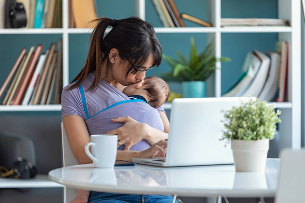 Tiro de muito jovem mãe beijando seu bebê em funda enquanto trabalhava com laptop em casa
. - Foto, Imagem