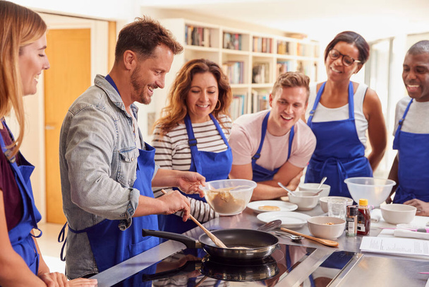 Male Teacher Making Pancake On Cooker In Cookery Class As Adult Students Look On - Foto, Bild