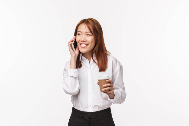 Office work, people and business concept. Portrait of cute asian girl drinking coffee and talking on phone with pleased, amused smile, having carefree conversation while on break, white background - Фото, изображение