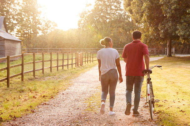 Rear View Shot Of Romantic Couple Walking And Pushing Bike Along Country Lane At Sunset - Foto, Imagem