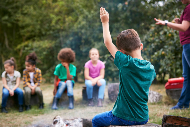 Children On Outdoor Activity Camping Trip Sit Around Camp Fire With Arms Raised Answering Question - Photo, image