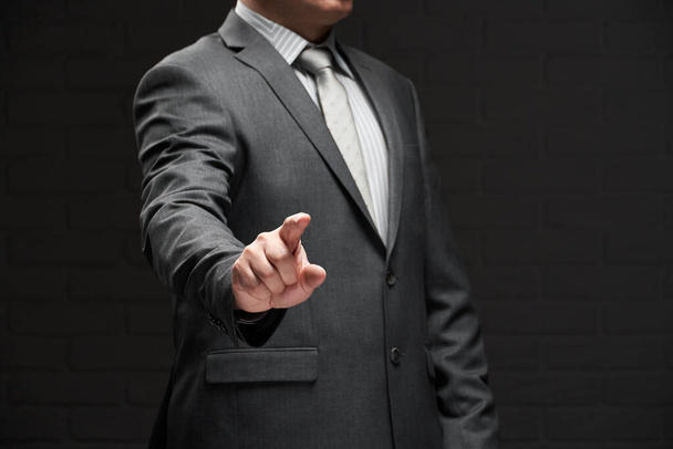 businessman portrait pointing by forefinger, dressed in gray suit, dark wall background - Foto, Imagem