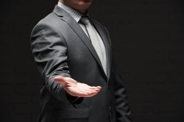 obchodník portrét ukazující dlaň ruky, demonstrovat něco, oblečený v šedém obleku, tmavé stěny pozadí - Fotografie, Obrázek