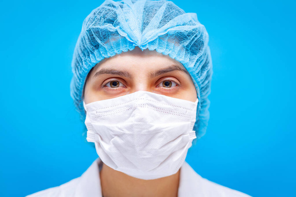 Γυναίκα γιατρός στην ιατρική μάσκα, καπέλο μαλλιών και λευκό φόρεμα portaiture σε μπλε φόντο κοντινό πλάνο. Ρηχό βάθος πεδίου. Ιός του Coronavirus, πανδημία και ιατρική περίθαλψη. - Φωτογραφία, εικόνα