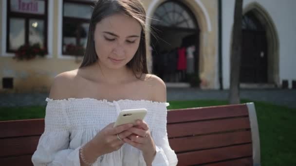 pretty woman is typing sms in cell phone sitting outdoors - Video, Çekim
