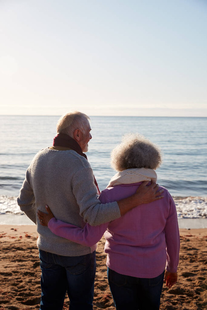 Rückansicht des liebenden Rentnerehepaares, das sich im Winter im Strandurlaub im Stehen umarmt - Foto, Bild