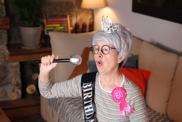 Nahaufnahme Porträt einer reifen Frau mit Diadem, Geburtstagsstreifen und 60 Jahre Geburtstagspreis, die während der Quarantäne zu Hause sitzt - Foto, Bild