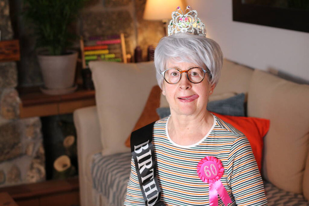 Nahaufnahme Porträt einer reifen Frau mit Diadem, Geburtstagsstreifen und 60 Jahre Geburtstagspreis, die während der Quarantäne zu Hause sitzt - Foto, Bild