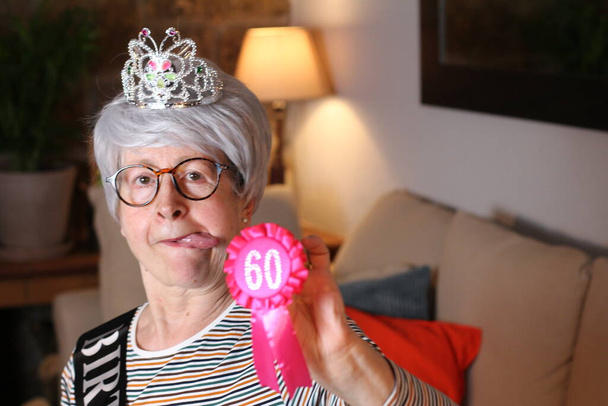 close-up portret van volwassen vrouw met tiara, verjaardagsstreep en 60 jaar verjaardagsprijs zittend thuis tijdens quarantaine - Foto, afbeelding