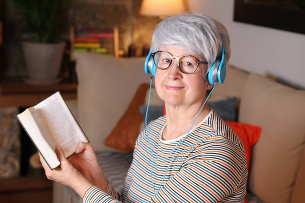 κοντινό πορτραίτο ώριμης γυναίκας που διαβάζει βιβλίο και ακούει μουσική με ακουστικά ενώ κάθεται στο σπίτι κατά τη διάρκεια της καραντίνας - Φωτογραφία, εικόνα