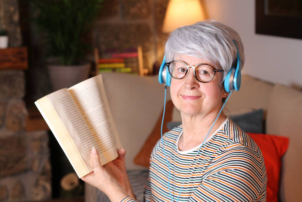 close-up portrait of mature woman reading book and listening music with headphone while sitting at home during quarantine - Photo, Image