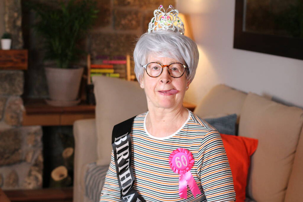 ritratto ravvicinato di donna matura con tiara, striscia di compleanno e premio di 60 anni seduta a casa durante la quarantena
 - Foto, immagini