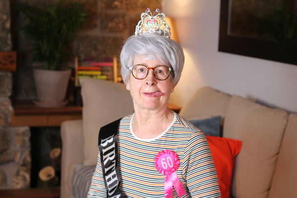 close-up portrait of mature woman with tiara, birthday stripe and 60 years birthday award sitting at home during quarantine - Photo, Image