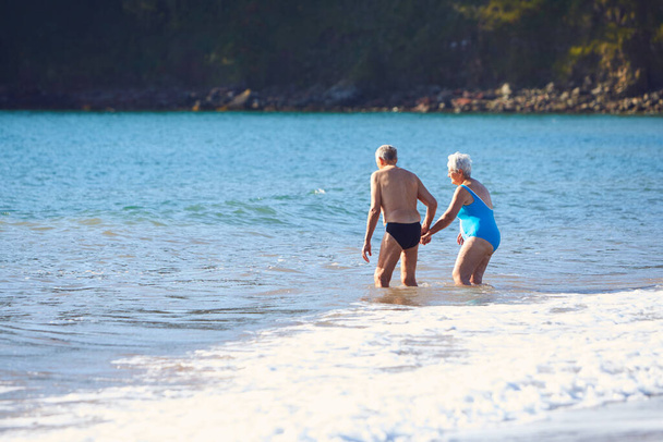 Loving Senior Couple Holding Hands As They Walk Into Sea For Swim - Photo, Image