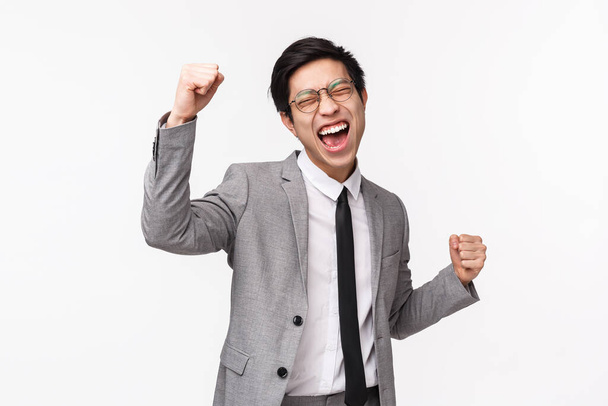 Waist-up portrait of relieved, successful asian young businessman, male entrepreneur in gray suit, fist pump, do champion dance, shouting yes victory, triumphing over achievement, winning - Photo, Image