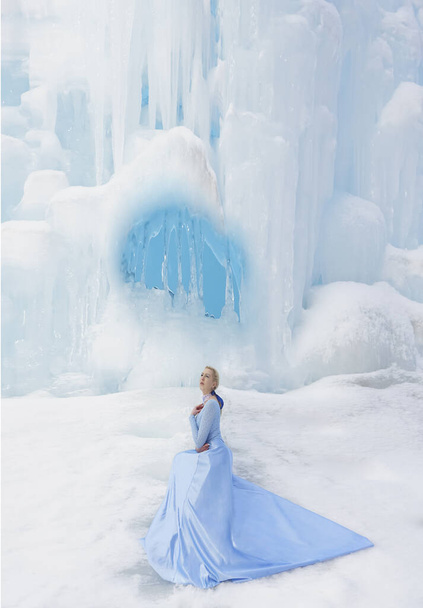 winter fairy tale on an ice waterfall a beautiful girl with long blonde hair and a blue dress with a long train - Φωτογραφία, εικόνα