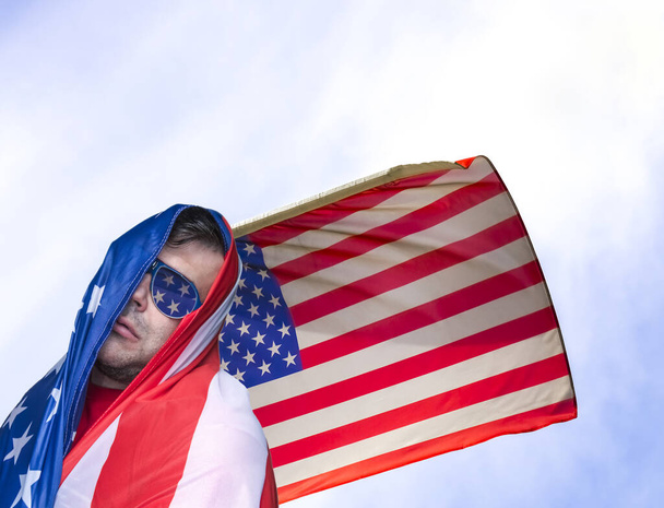 Independence day of United States of America. Adult Man wrapped in American Flag stand against cloudy blue sky background. young guy wrapped in  USA flag on heaven backdrop.  - 写真・画像
