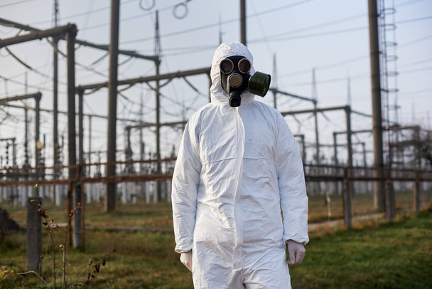 Scientist wearing white protective uniform, gas mask and gloves on the territory of thermal power station, power lines on background - Photo, Image