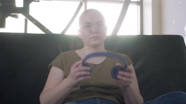 Lesbian bald girl on the couch in professional headphones. Listens to music, relaxes at home. Home rest. Rehabilitation after chemotherapy. - Felvétel, videó
