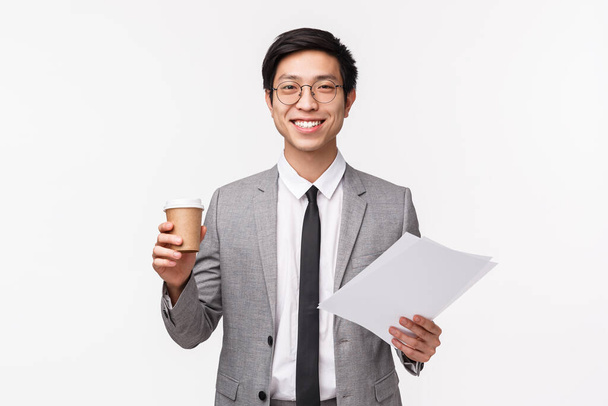 Office workers, business and lifestyle concept. Waist-up portrait of upbeat, enthusiastic young asian businessman drinking morning coffee at office, smiling pleased as holding documents - Foto, Bild