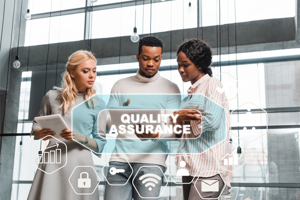 young businesswoman holding digital tablet while standing near african american colleagues looking at documents, quality assurance illustration - Photo, Image