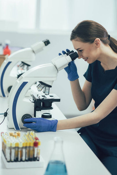 Cheerful lab assistant sitting in front of a microscope and looking at slides - Фото, изображение