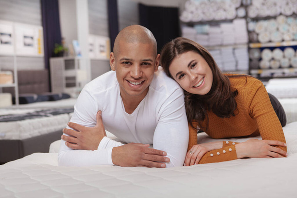 Happy young couple smiling to the camera, lying on orthopedic mattress at furniture store. Buying new bed concept - 写真・画像