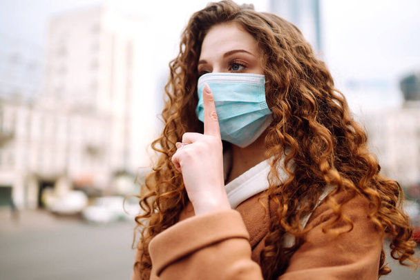 Girl in protective sterile medical mask on her face shows a gesture: shh. silence. The concept of preventing the spread of the epidemic and treating coronavirus, pandemic in quarantine city.  - Фото, изображение