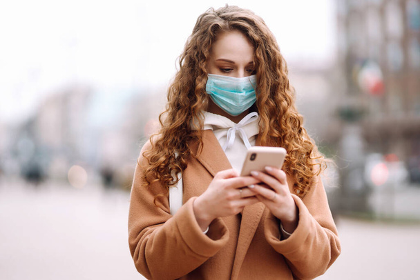 Girl in protective sterile medical mask on her face with a phone in  quarantine city. Woman using the phone to search for news. The concept of preventing the spread of the epidemic. - Фото, изображение