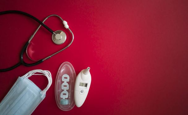 Stetoscopio, confezione di maschere mediche, termometro auricolare elettronico con set di attacchi monouso in contenitore disteso su fondo rosso brillante a sinistra.Copia spazio.Prevenzione e diagnosi del coronavirus
 - Foto, immagini