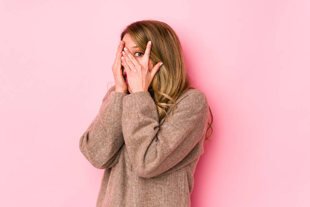 Νεαρή λευκή γυναίκα απομονωμένη σε ροζ φόντο αναβοσβήνει μέσα από τα δάχτυλα φοβισμένη και νευρική. - Φωτογραφία, εικόνα