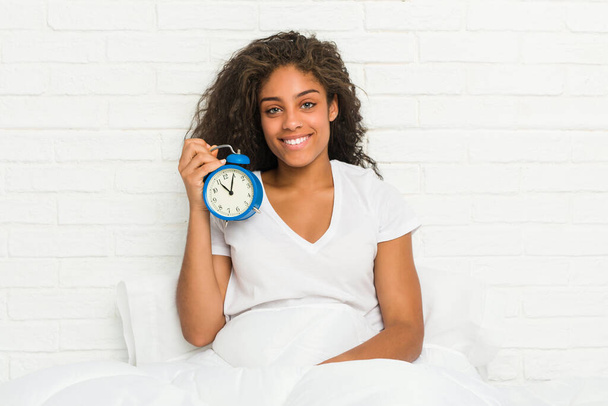 Молодая африканская американка сидит на кровати, держа будильник счастливым, улыбающимся и веселым
. - Фото, изображение