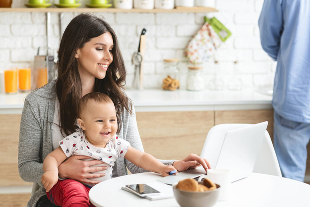Millennial moeder werken op laptop met baby in de keuken - Foto, afbeelding