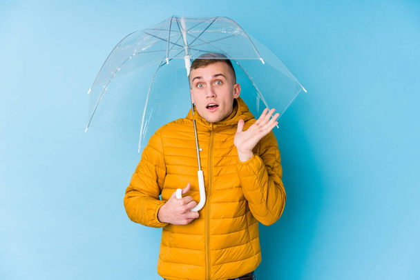 Молодой кавказский мужчина с зонтиком удивлен и шокирован
. - Фото, изображение
