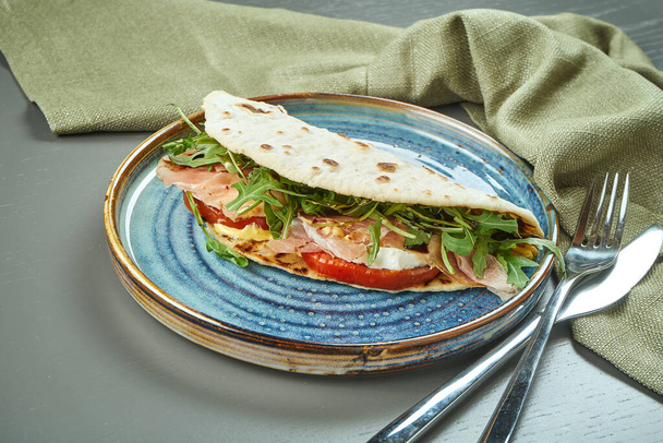 Piadina - tortilla-pão italiano clássico com prosciutto, arugula, mussarela e tomates em uma chapa azul na mesa de madeira. Sanduíche rápido
 - Foto, Imagem