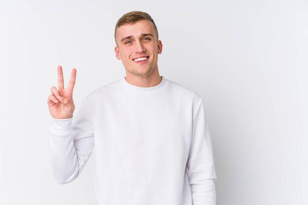 Jonge blanke man op witte achtergrond met overwinningsteken en brede glimlach. - Foto, afbeelding